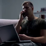 Frustrated veteran sitting on his couch with his laptop talking on the phone to the VA about his disability benefits